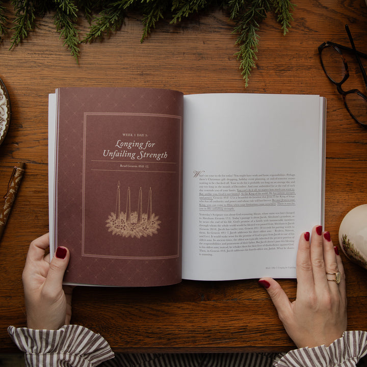 An open book held by two hands with red painted fingernails, placed on a wooden table. The left page shows a brown cover with the title 'Longing for Unfailing Strength' and a candle illustration, while the right page contains printed text.