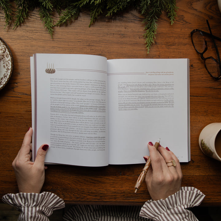 A top-down view of a person's hands holding an open book on a wooden table, with a pen in one hand and holiday greenery and glasses around it.