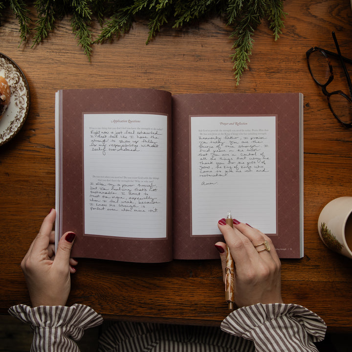 An open journal with handwritten pages on a rustic wooden table, held by two hands. The scene is styled with pine branches, a plate, eyeglasses, and a mug.