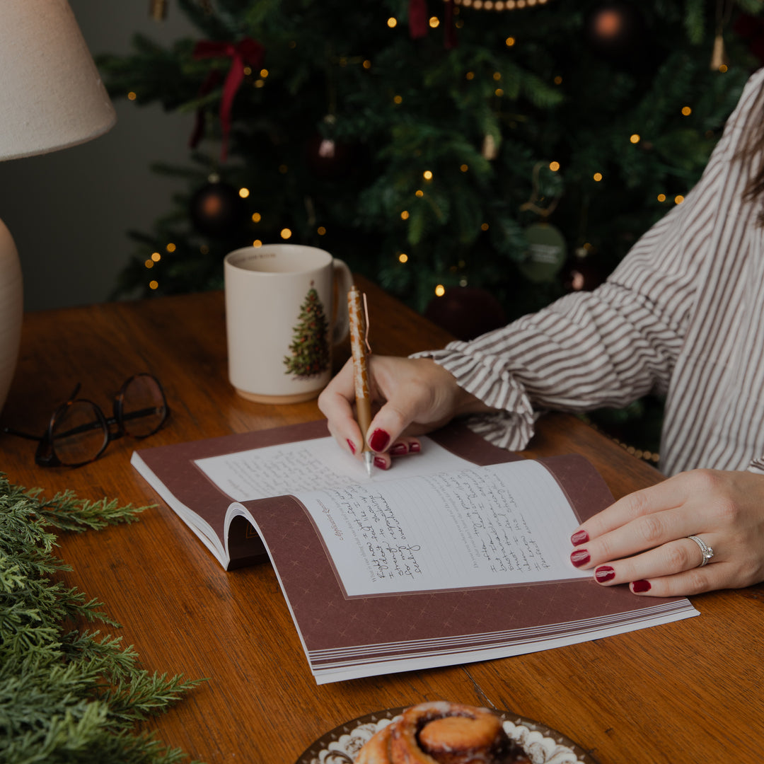 A person writing in a decorative book or journal on a wooden table, with a Christmas tree and warm holiday lights in the background. A white mug with a green tree design sits nearby, along with eyeglasses and a pastry.
