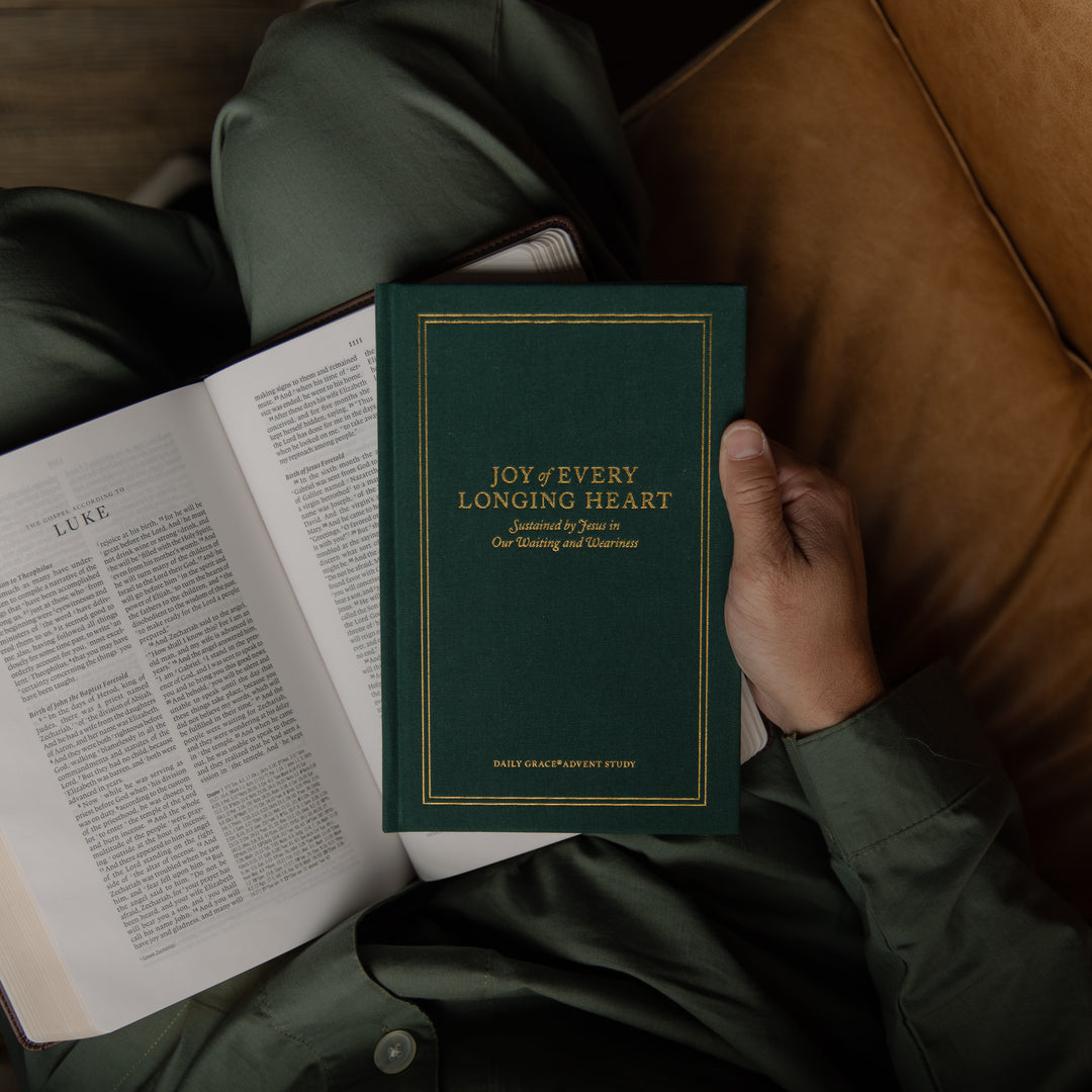 A person in dark green pajamas holds a dark green book titled 'Joy of Every Longing Heart' next to an open Bible on a brown leather couch.