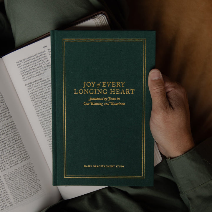 A person in dark green pajamas holds a dark green book titled 'Joy of Every Longing Heart' next to an open Bible on a brown leather couch.