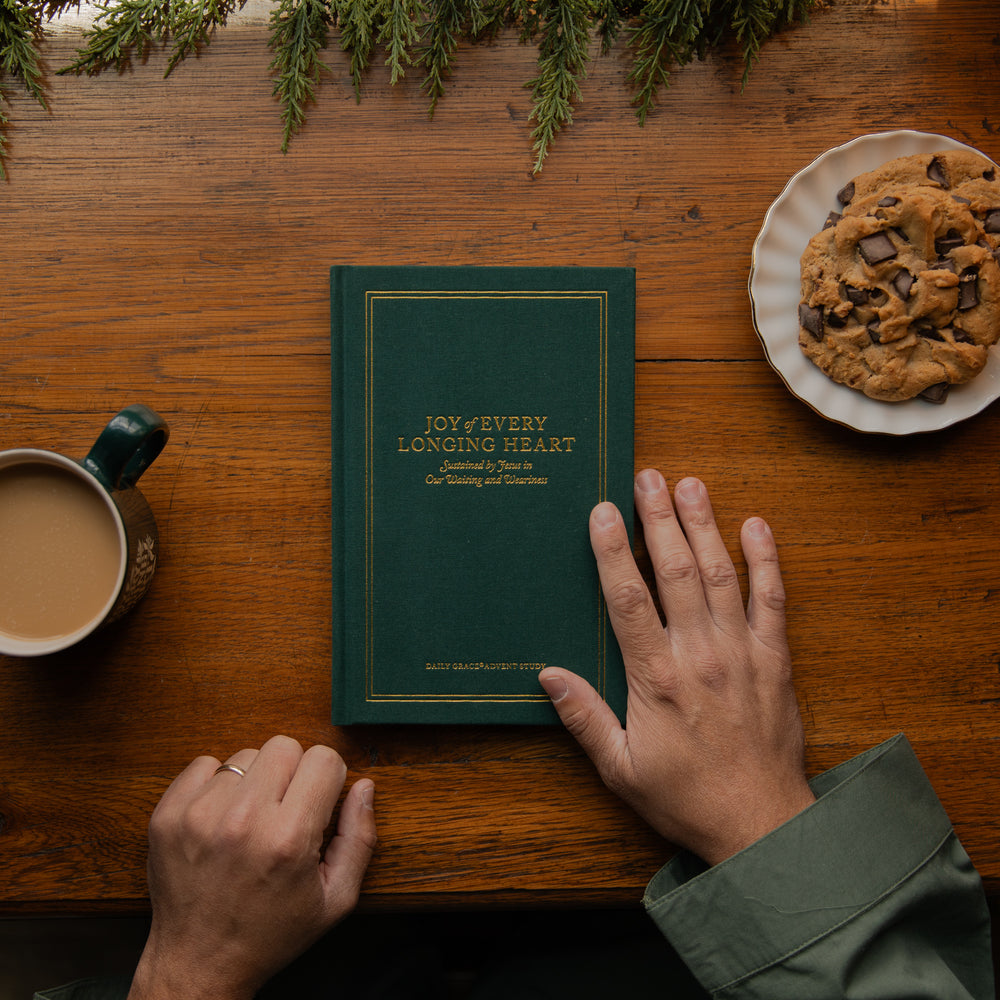 A dark green hardcover book titled 'JOY & EVERY LONGING HEART' lies on a wooden table, with two hands resting near it. A cup of coffee, a plate of chocolate chip cookies, and evergreen branches are visible around it in a cozy tabletop setting.
