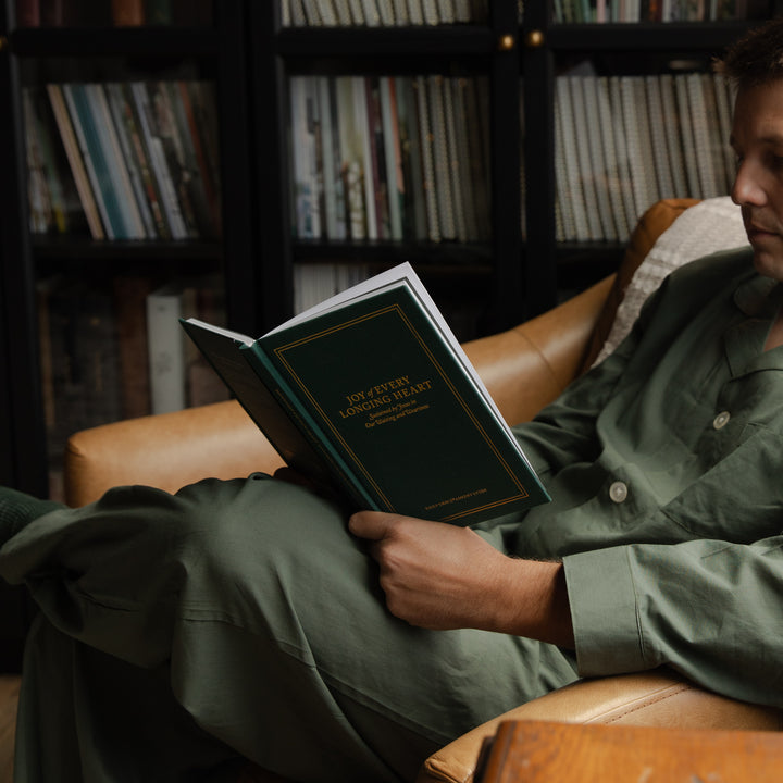 A person in olive green pajamas sits in a tan leather armchair, holding an open hardcover book titled 'How to Build a Business Like Donald Trump' in front of a dark bookshelf.