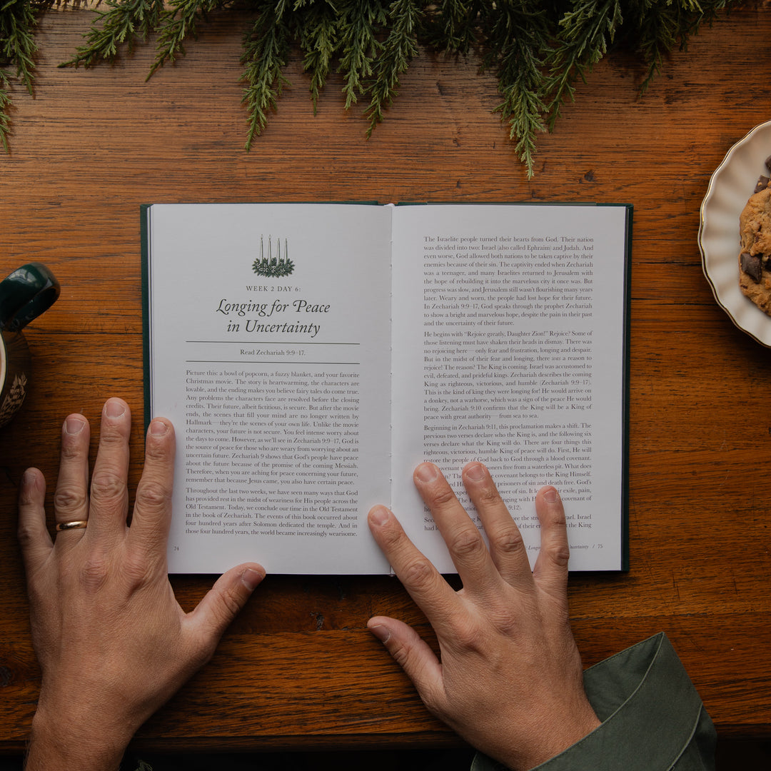 An open book titled 'Waiting for Peace in Uncertainty' resting on a wooden table, with two hands holding it open. Evergreen branches and a plate of cookies are visible in the background and surroundings.