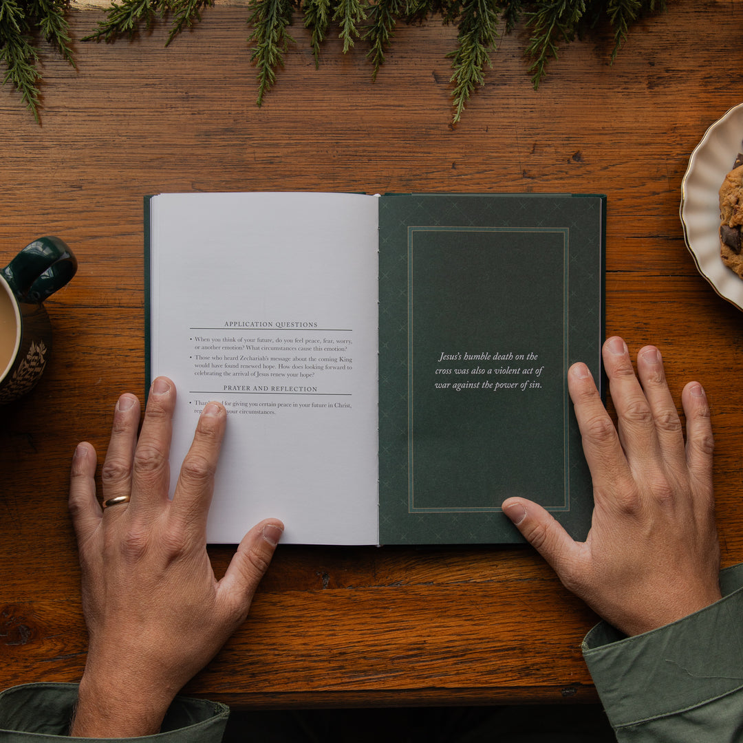 An open book lying on a wooden table, with two hands holding the pages. The left page is white with printed text, and the right page is dark green with a bordered text box containing a quote.