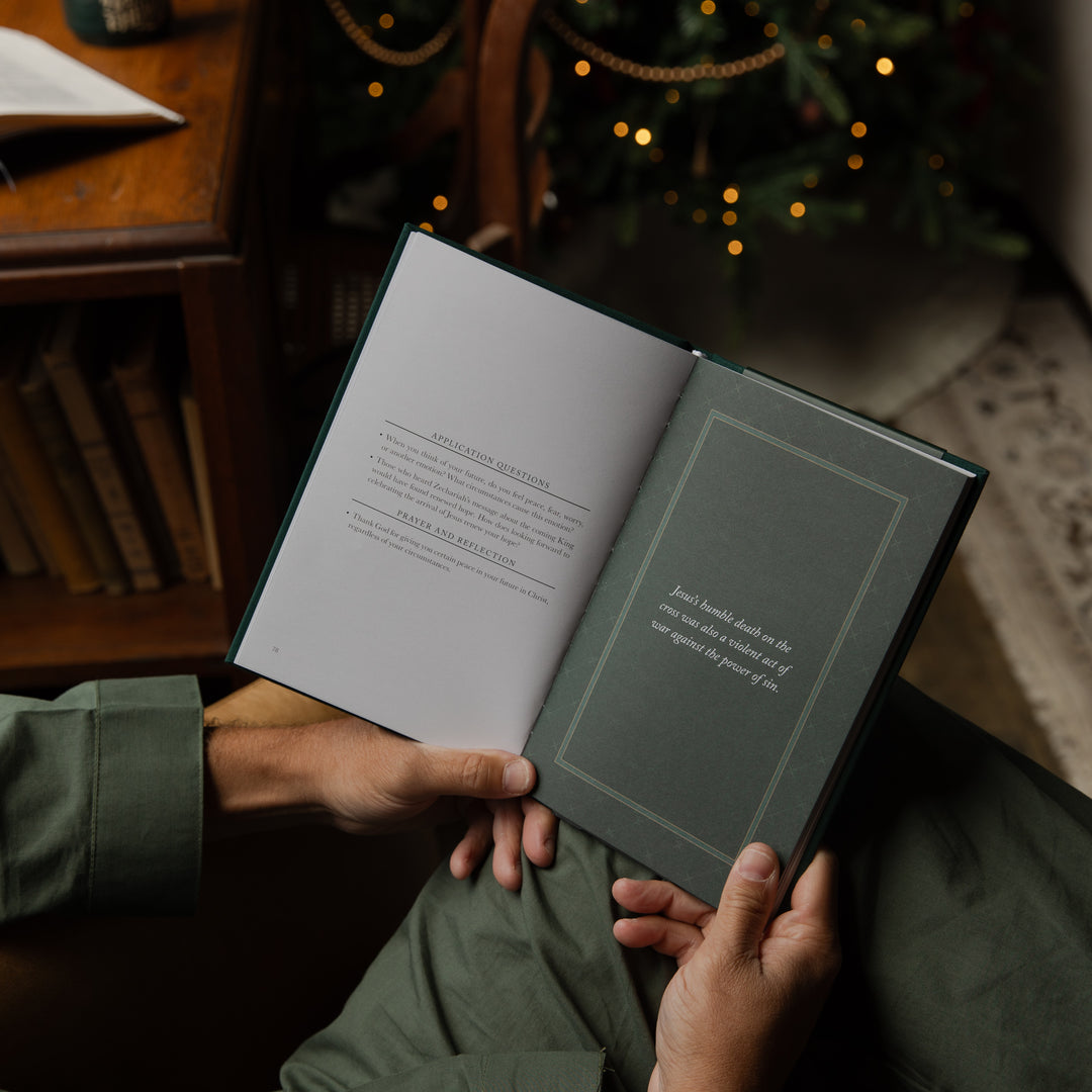 A person holding an open book in a cozy indoor setting, with a blurred Christmas tree and warm lights in the background. The book has a dark green cover and open pages with printed text.