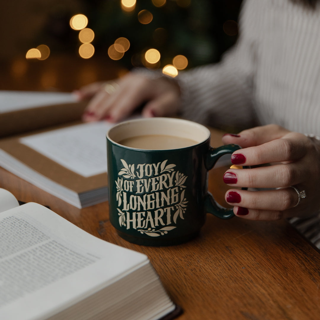 A woman with her hand on the handle of a green mug that says Joy of Every Longing Heart on a brown wooden desk