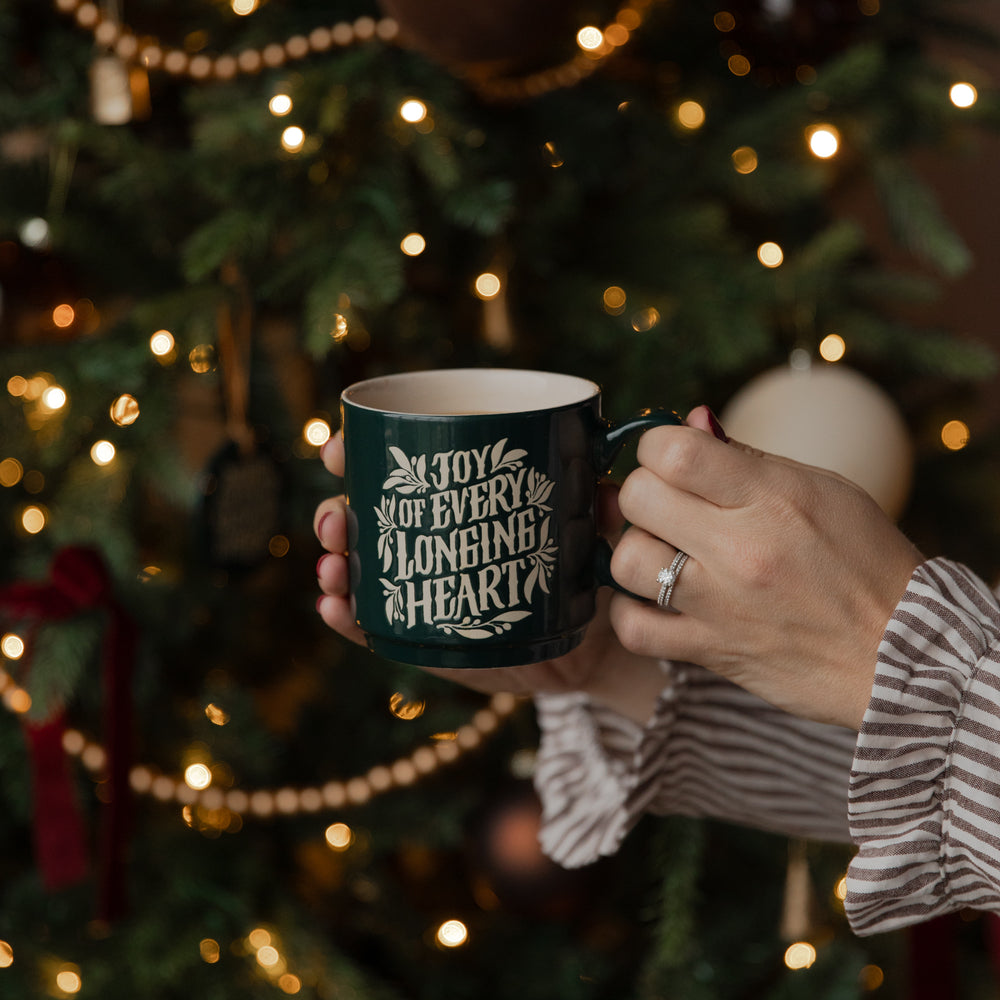 A person holds a dark green ceramic mug with white text reading 'JOY OF EVERY 2 LONGING 2 HEARTS' in front of a blurred Christmas tree with warm lights.