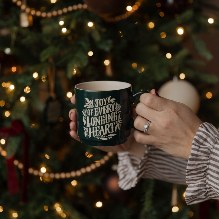 A person holds a dark green ceramic mug with white text reading 'JOY OF EVERY 2 LONGING 2 HEARTS' in front of a blurred Christmas tree with warm lights.