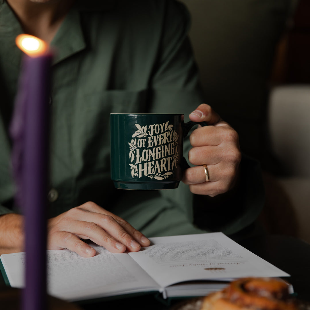 A person in a dark green shirt holds a dark green ceramic mug with gold lettering that reads 'IN JOY OF EVERY LONGING IS A HEART'. The scene is warmly lit by a purple candle in the foreground, with an open book and a pastry on a table below.