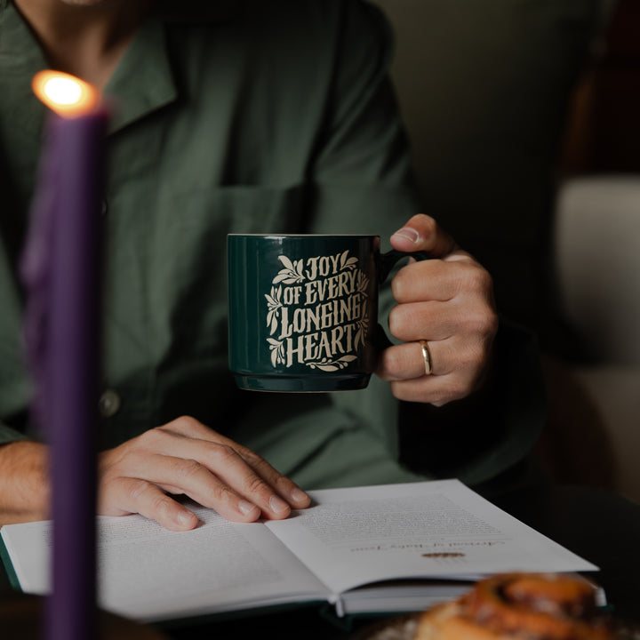 A person in a dark green shirt holds a dark green ceramic mug with gold lettering that reads 'IN JOY OF EVERY LONGING IS A HEART'. The scene is warmly lit by a purple candle in the foreground, with an open book and a pastry on a table below.