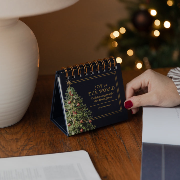 A small spiral-bound desk flip book with a Christmas tree illustration and the text 'JOY to THE WORLD' stands on a wooden table. A person's hand with red nail polish is touching it, with a lamp on the left and a blurred Christmas tree in the background.