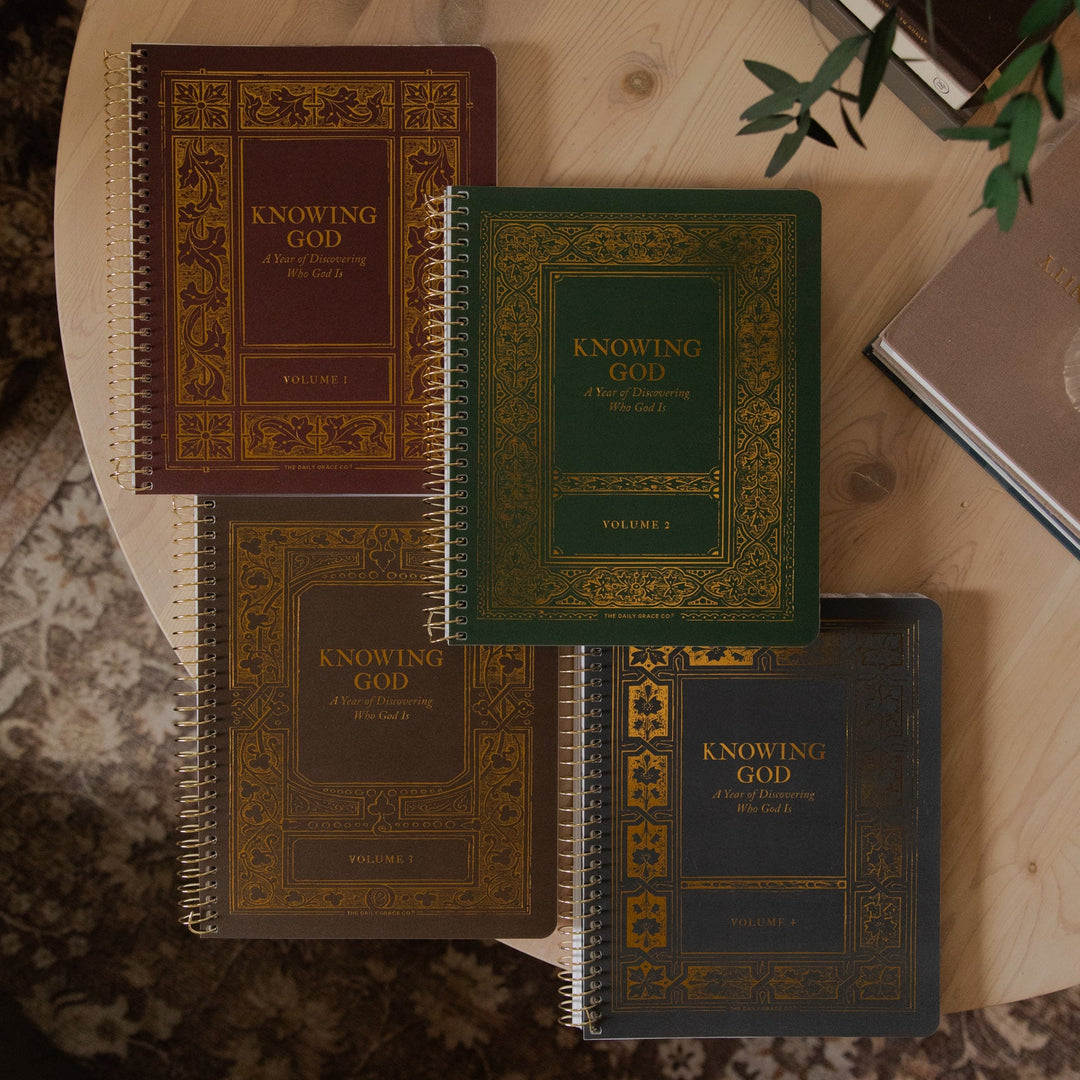 Four spiral-bound books titled 'Knowing God' in different colors (burgundy, green, tan, and navy) arranged on a round wooden table with a glass and greenery in the background.