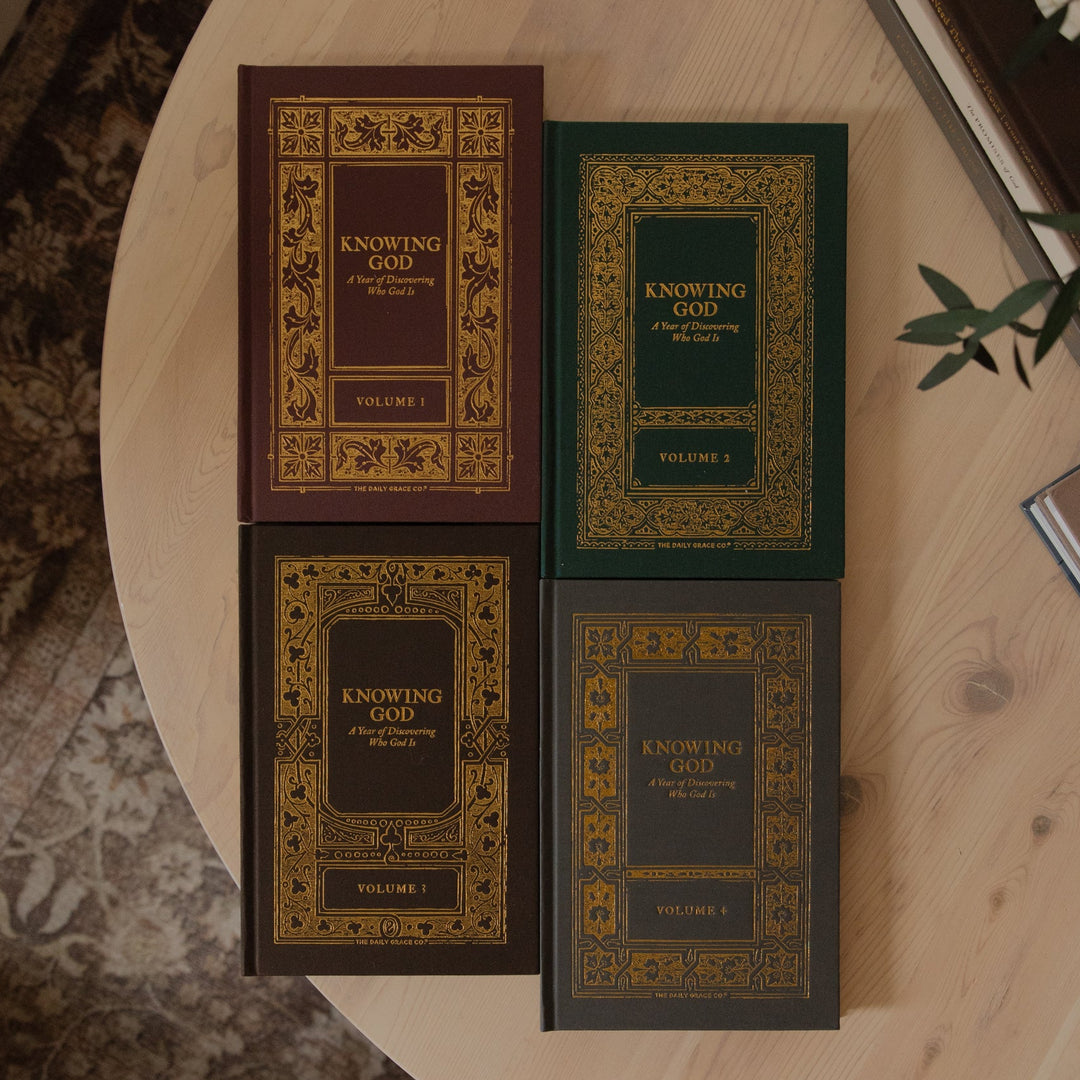 Four hardcover books titled 'Knowing God' arranged on a round wooden table, with two in dark brown and two in dark green, each featuring ornate gold decorative borders and gold lettering. The books are styled in a cozy home environment with a patterned rug, a plant, and other books visible.