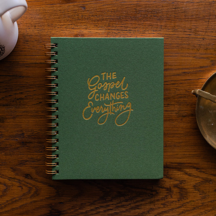 The Gospel Changes Everything Not taking Journal on a wooden table next to a coffee cup and an bronze oil lamp