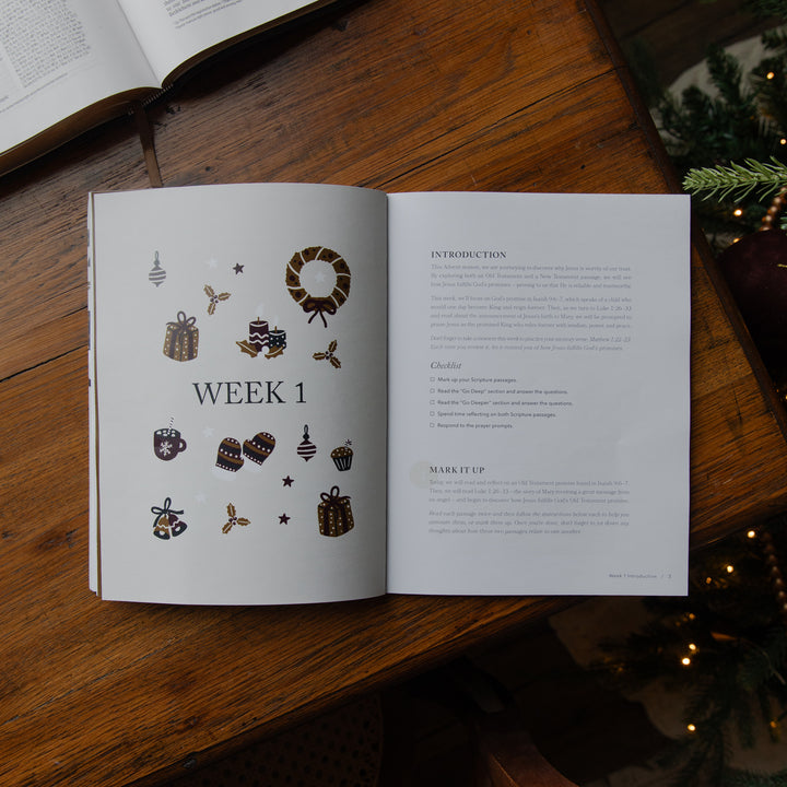 An open printed booklet or guide laid on a wooden table, showing a left page with small holiday illustrations and the heading 'WEEK 1' plus a right page with introduction and chapter text. An open book and Christmas decorations are visible around it.