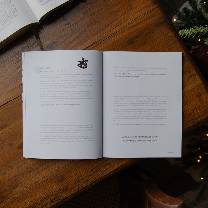An open book laid on a rustic wooden table, showing printed pages with text and a small illustration. The scene includes part of another open book, a chair edge, and evergreen branches with warm string lights in the background.