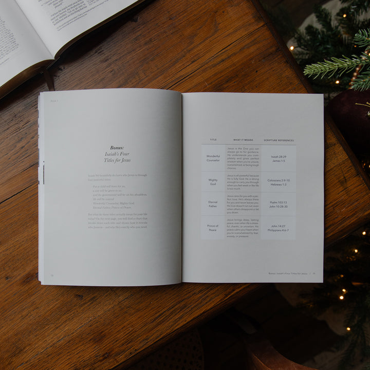 An open book laid flat on a wooden table, showing printed text pages. The scene includes part of another open book at the top left and evergreen branches with warm lights at the right edge, creating a styled tabletop setting.