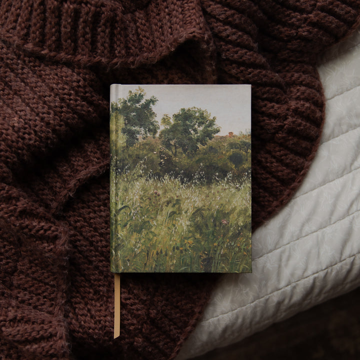 A hardcover notebook featuring an impressionist landscape painting of a green grassy field and trees, styled on a dark brown chunky knit blanket next to a white quilted pillow.