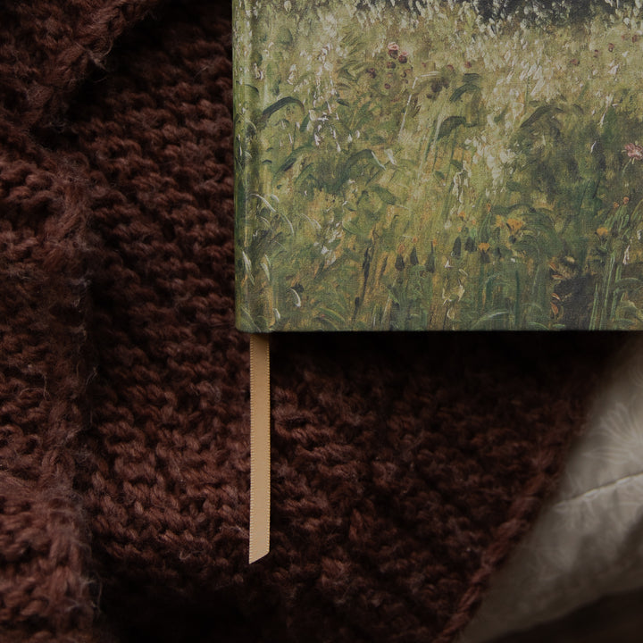 A close-up of a hardcover book with a textured green and yellow floral or grassy cover design, resting on a chunky dark brown knit blanket. A light beige ribbon bookmark is visible hanging from the book's edge.