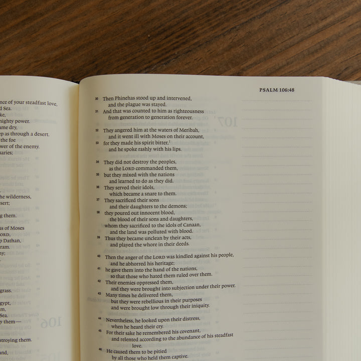 An open book showing printed text from Psalm 106:48 on both pages, placed on a dark wood surface.