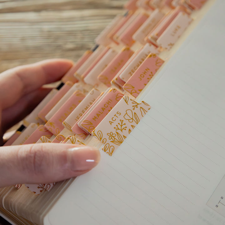 Hand holding a stack of pink and gold bible tabs with names on them, over a notebook.