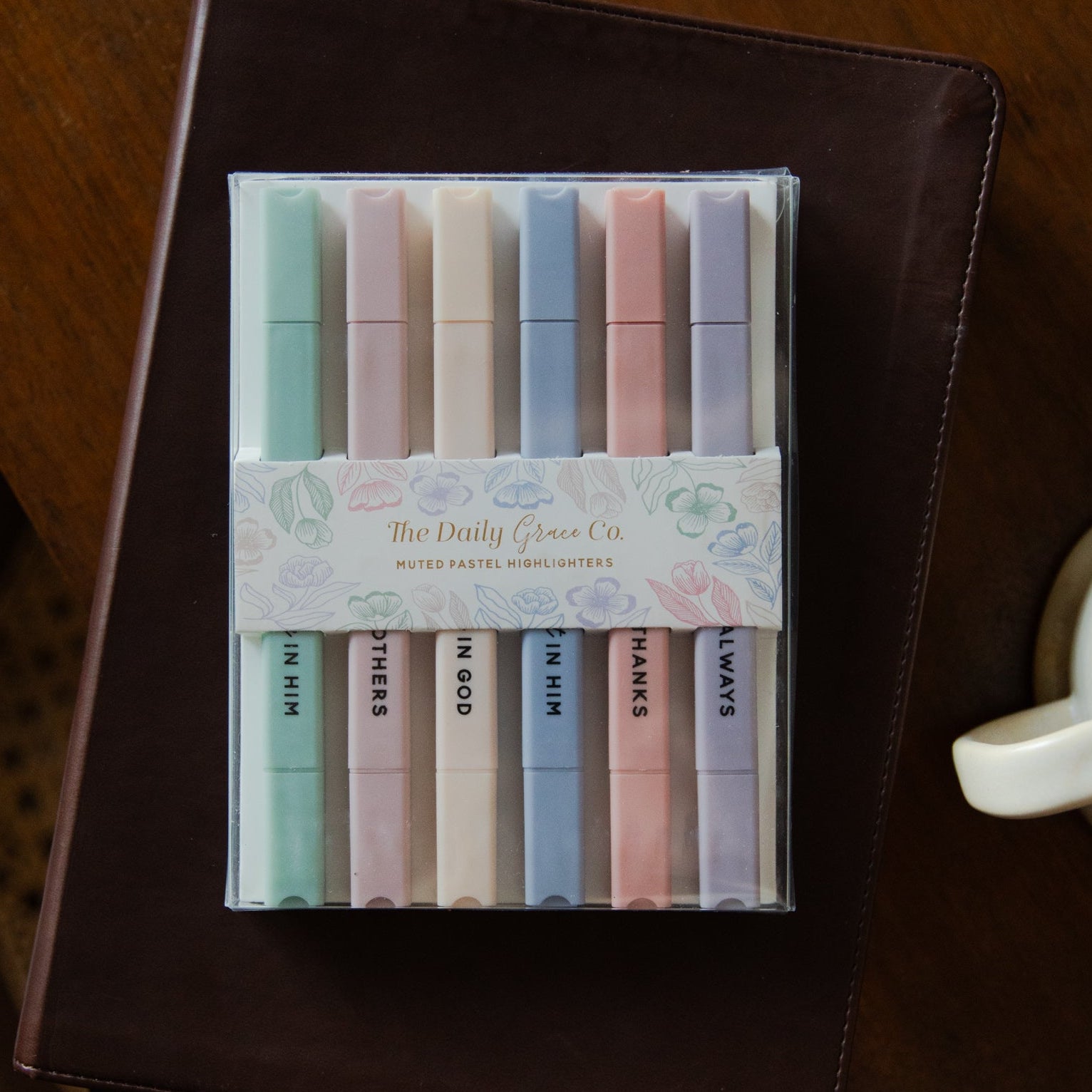 Set of pastel-colored markers in a clear plastic case on a wooden surface with a notebook and mug.