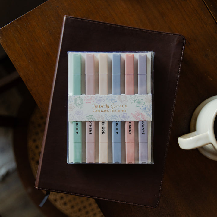 Set of pastel-colored markers in a clear plastic case on a wooden surface with a notebook and mug.