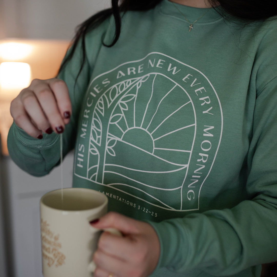 Woman wearing a green Christian sweatshirt that says His mercies are new every morning, grabbing a teabag out of a coffee cup