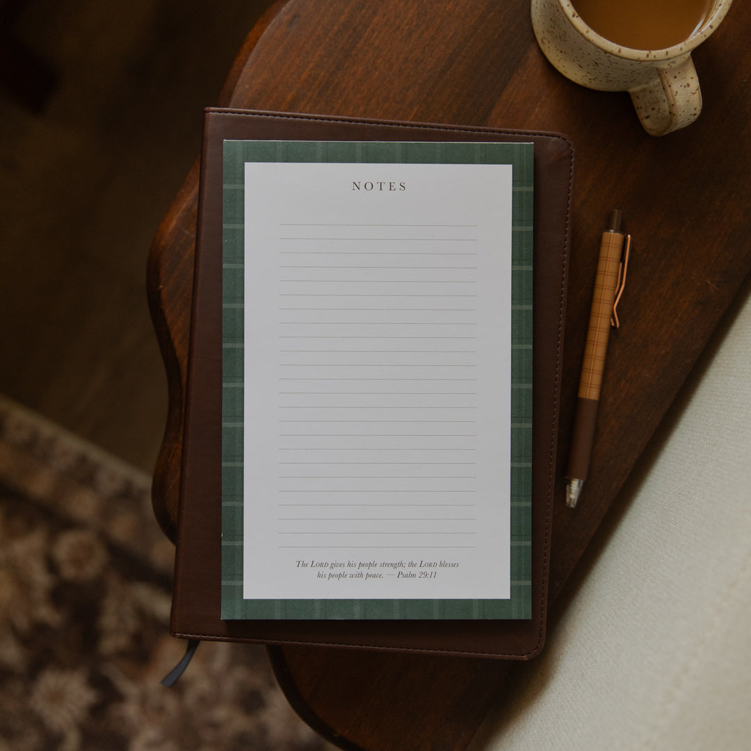 A brown leather-bound notebook with a green plaid patterned page protector and white lined notes paper, styled on a dark wooden table next to a pen and a ceramic mug.