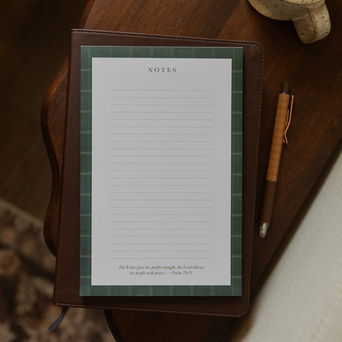 A brown leather-bound notebook with a green plaid patterned page protector and white lined notes paper, styled on a dark wooden table next to a pen and a ceramic mug.