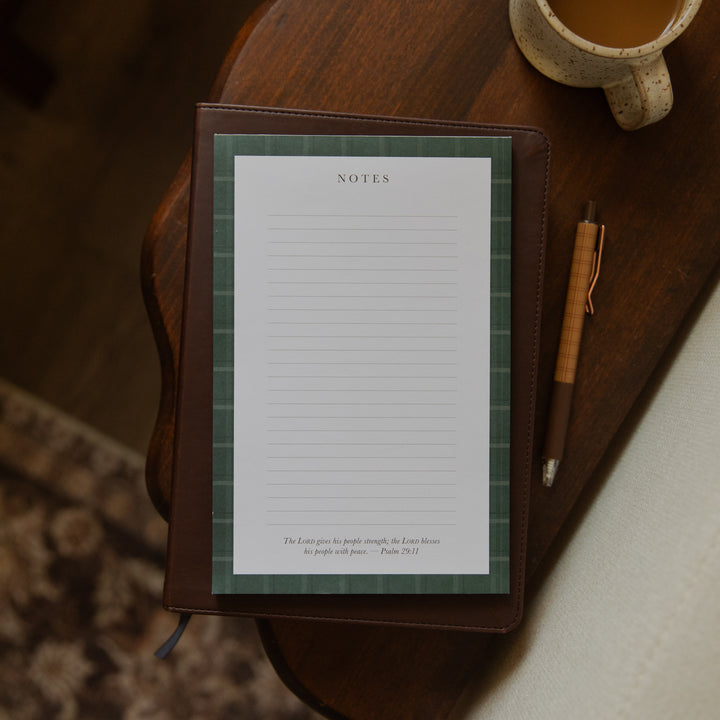 A brown leather-bound notebook with a green plaid patterned page protector and white lined notes paper, styled on a dark wooden table next to a pen and a ceramic mug.