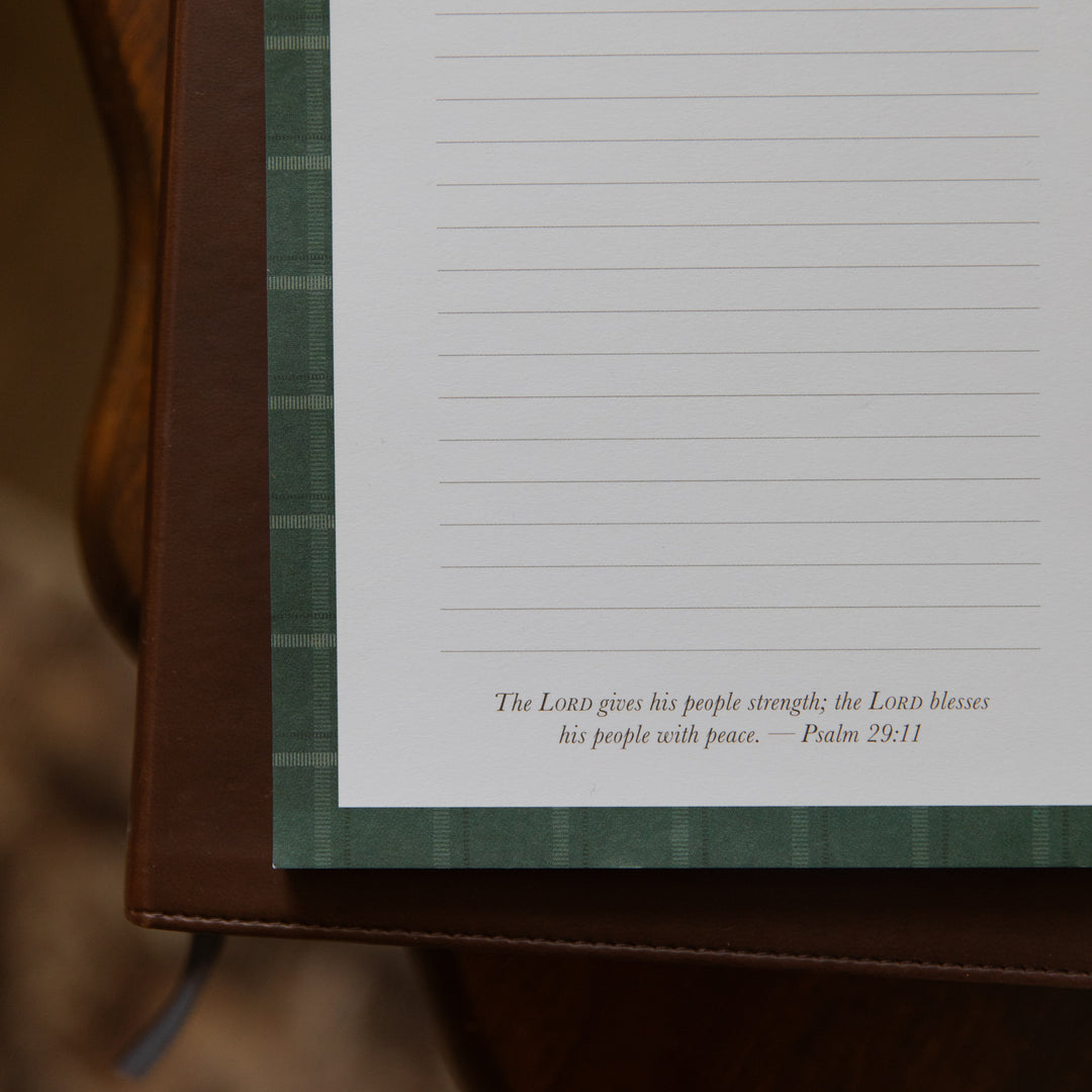 A close-up of a lined journal page with a green plaid border and a Bible verse printed at the bottom, resting on a brown leather-bound book or journal.