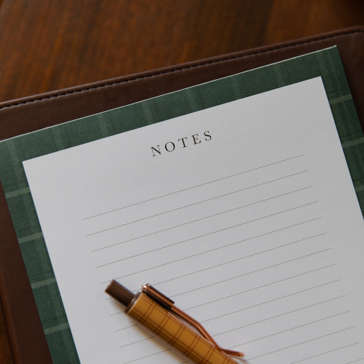 A close-up of a lined notes pad with a green plaid border, placed on a brown leather folio with a tan plaid pen resting on it, set on a wooden surface.