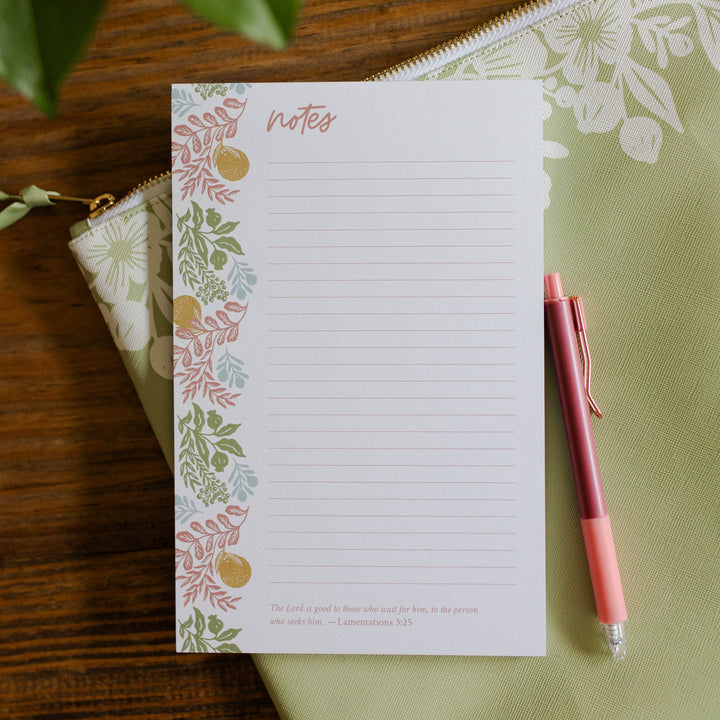 Image of a note pad with oranges and a verse at the bottom of the notepad. There is also a green pouch and coral pen. 