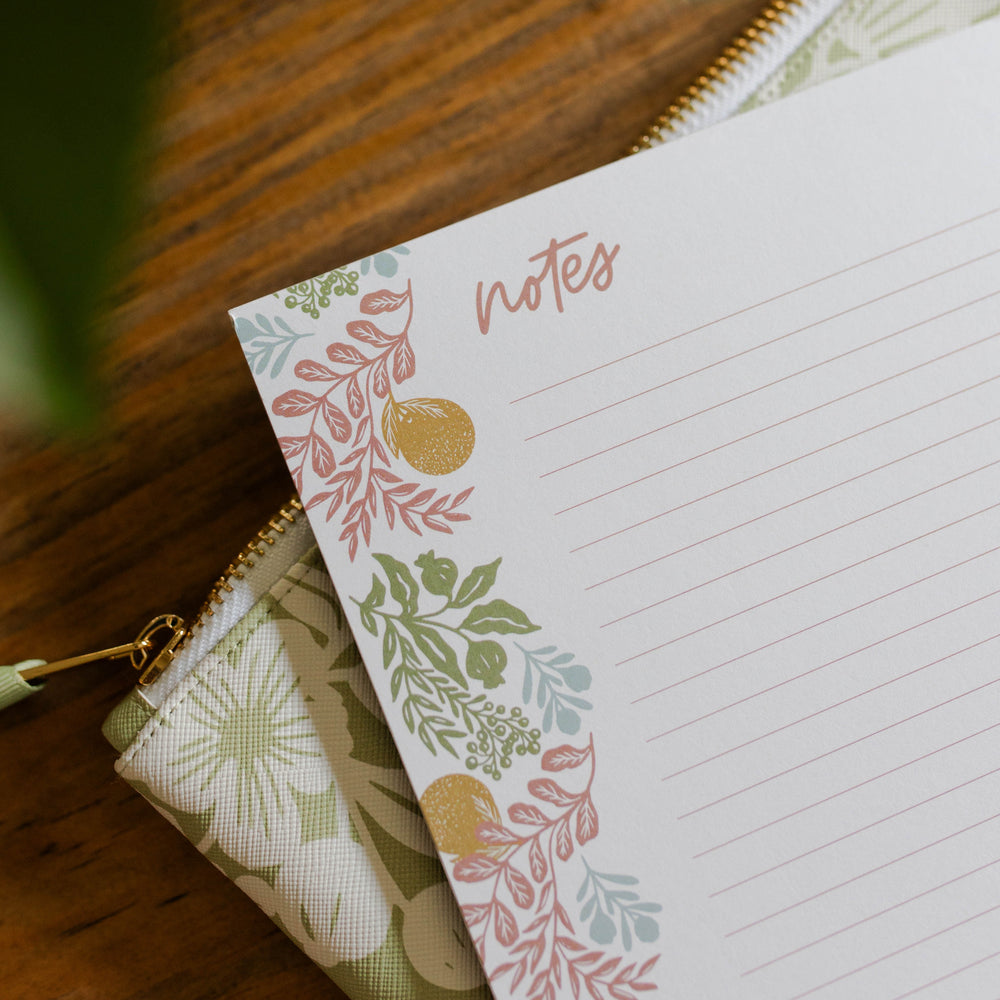 Image of a notepad that is coral, orange, green, and blue. The notepad is on top of a green, floral pouch. 