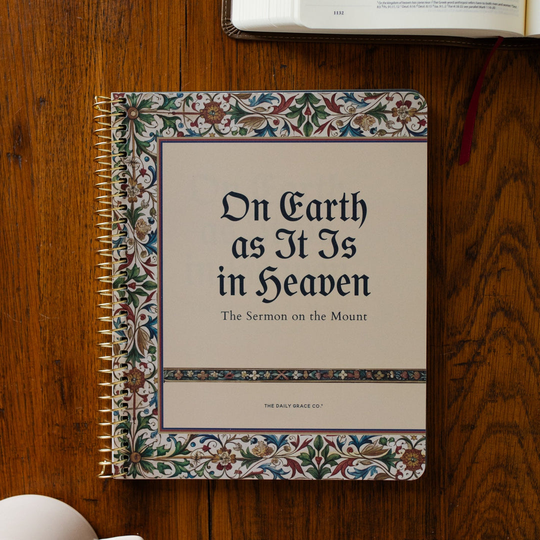 Notebook titled 'On Earth as It Is in Heaven' on a wooden surface with an open book and coffee cup.