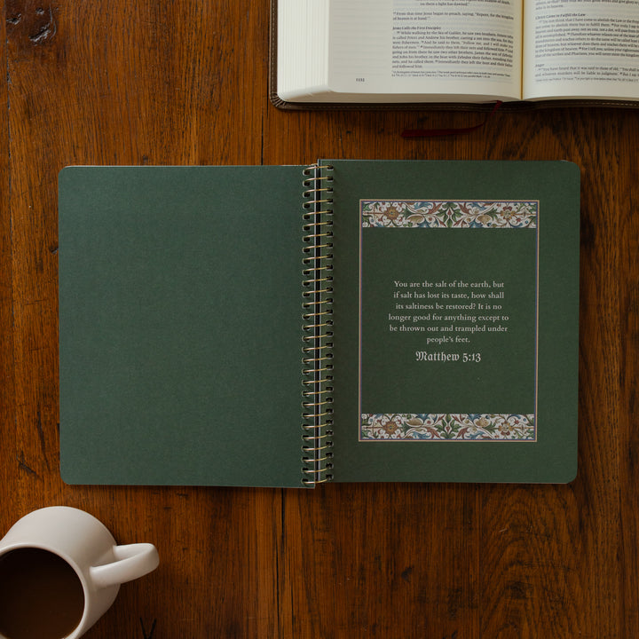 Open green notebook with text and decorative elements on a wooden surface, next to an open book and a white mug.