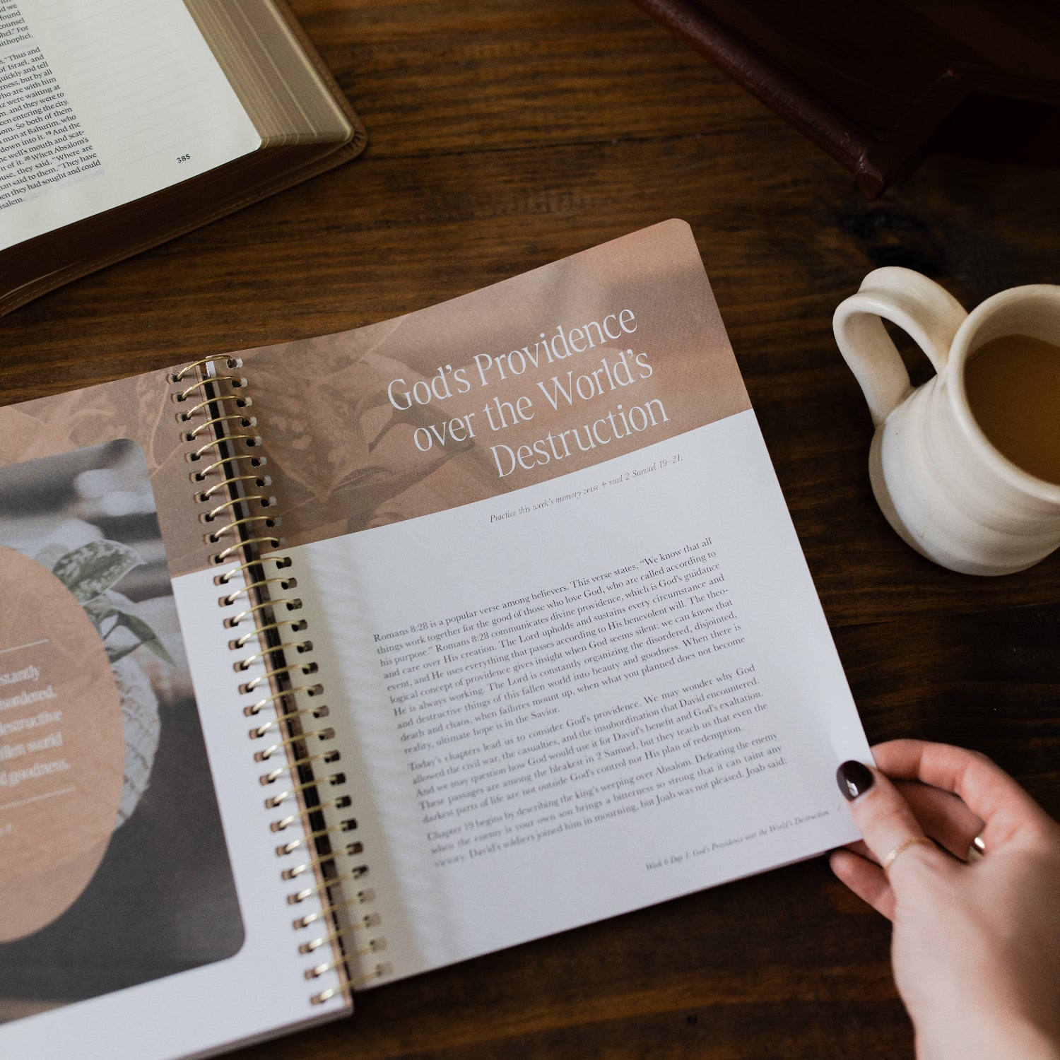 Peaks and Valleys Spiral Study opened to the Gods Providence page with a woman's hand on the corner next to a dark brown wooden tray and an open bible and a white coffee cup on a brown wooden table
