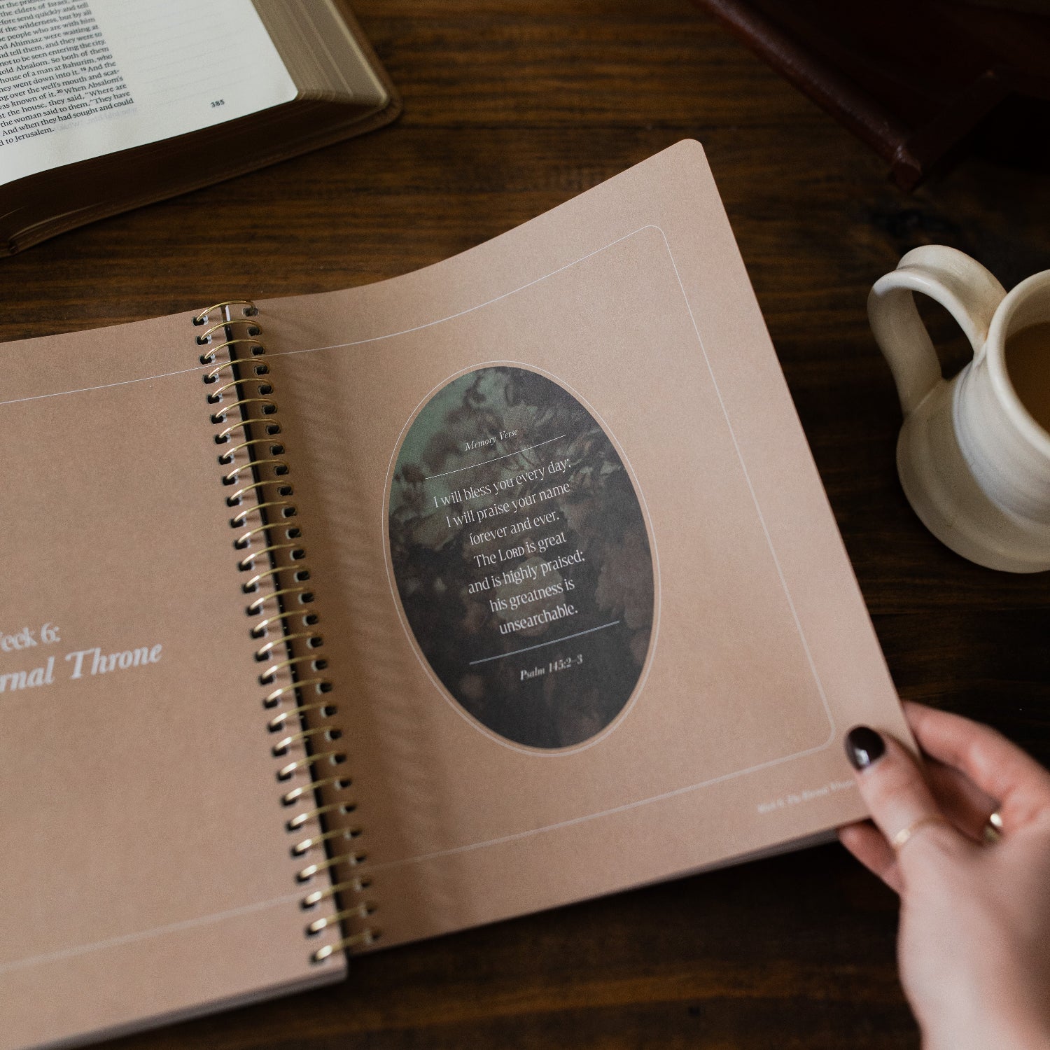 Peaks and Valleys Spiral Study opened to a memory verse page with a woman's hand holding the corner next to a dark brown wooden tray and an open bible and a white coffee cup on a brown wooden table 