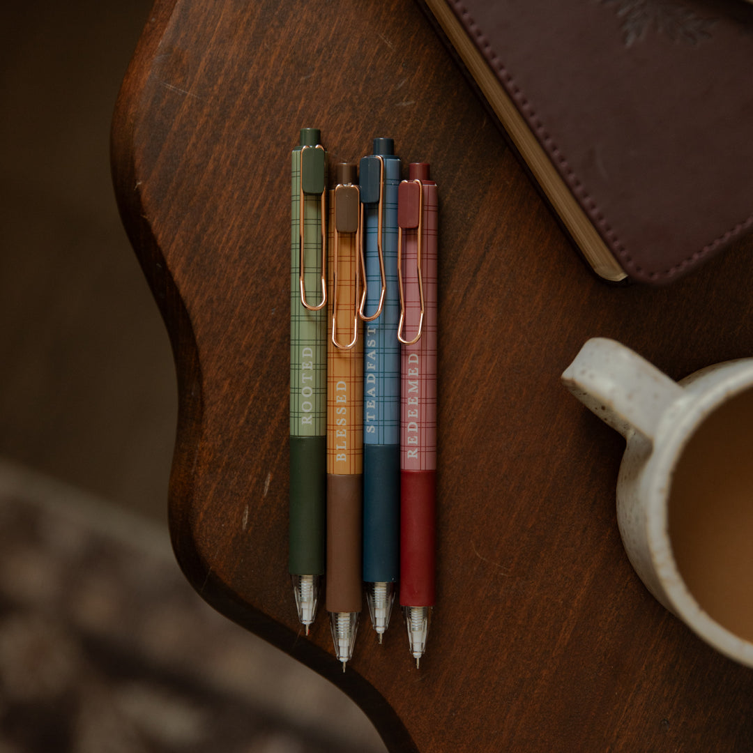 Four slim retractable pens in muted earthy colors are arranged on a dark wooden surface next to a notebook and a ceramic mug. The pens have visible clip details and vertical text on the barrels.