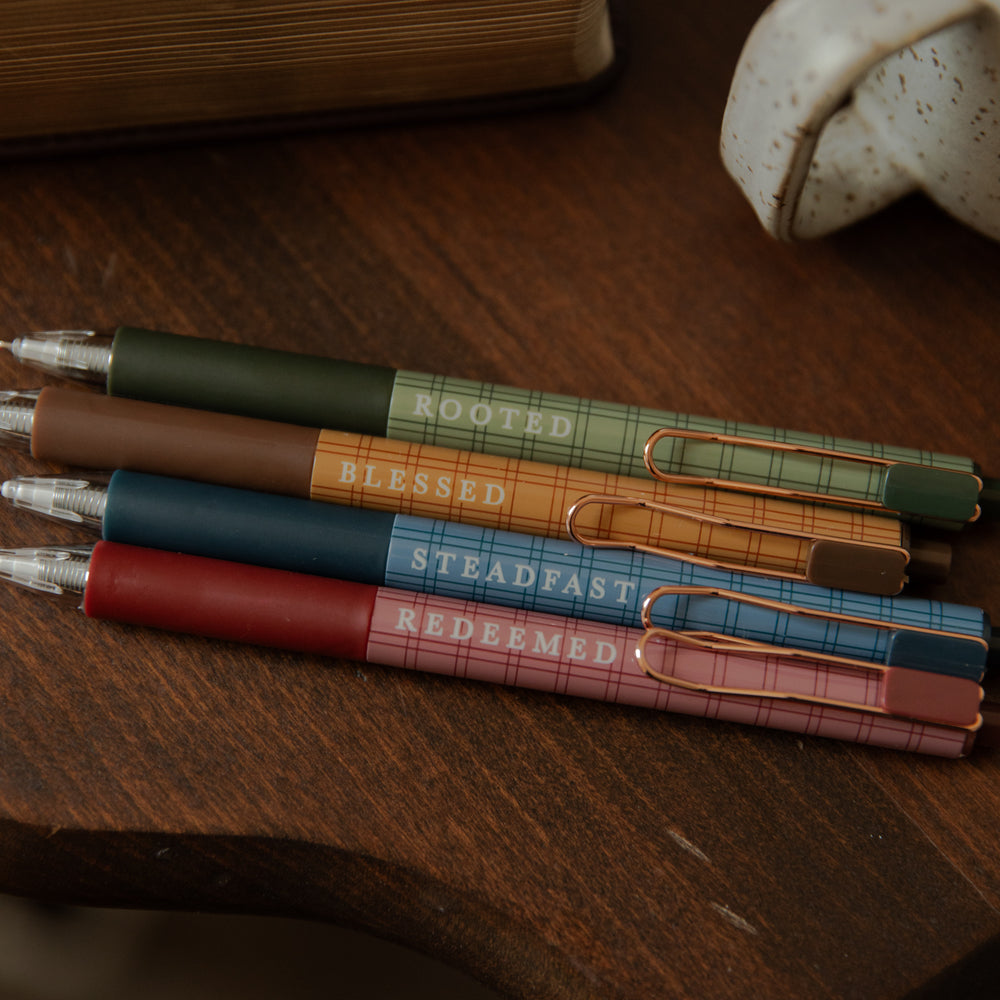 Four plaid retractable pens in green, brown, blue, and pink, stacked diagonally on a dark wooden table. Each pen has a different color body with a gold-tone pocket clip and is printed with a religious phrase: 'ROOTED', 'BLESSED', 'STEADFAST', and 'REDEEMED'.
