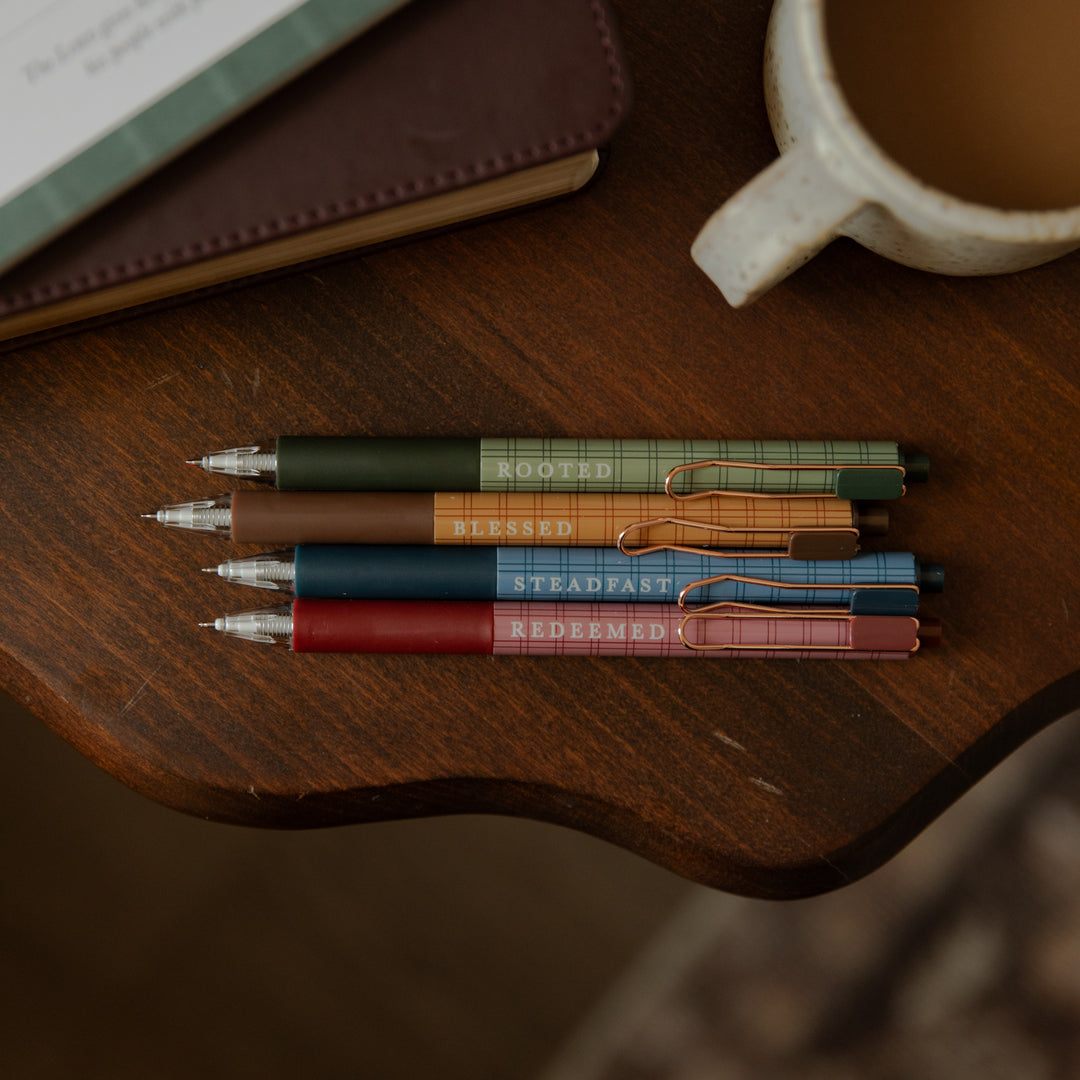 Four mechanical pencils with plaid-patterned barrels in green, brown, blue, and red, arranged on a dark wooden table next to a ceramic mug.