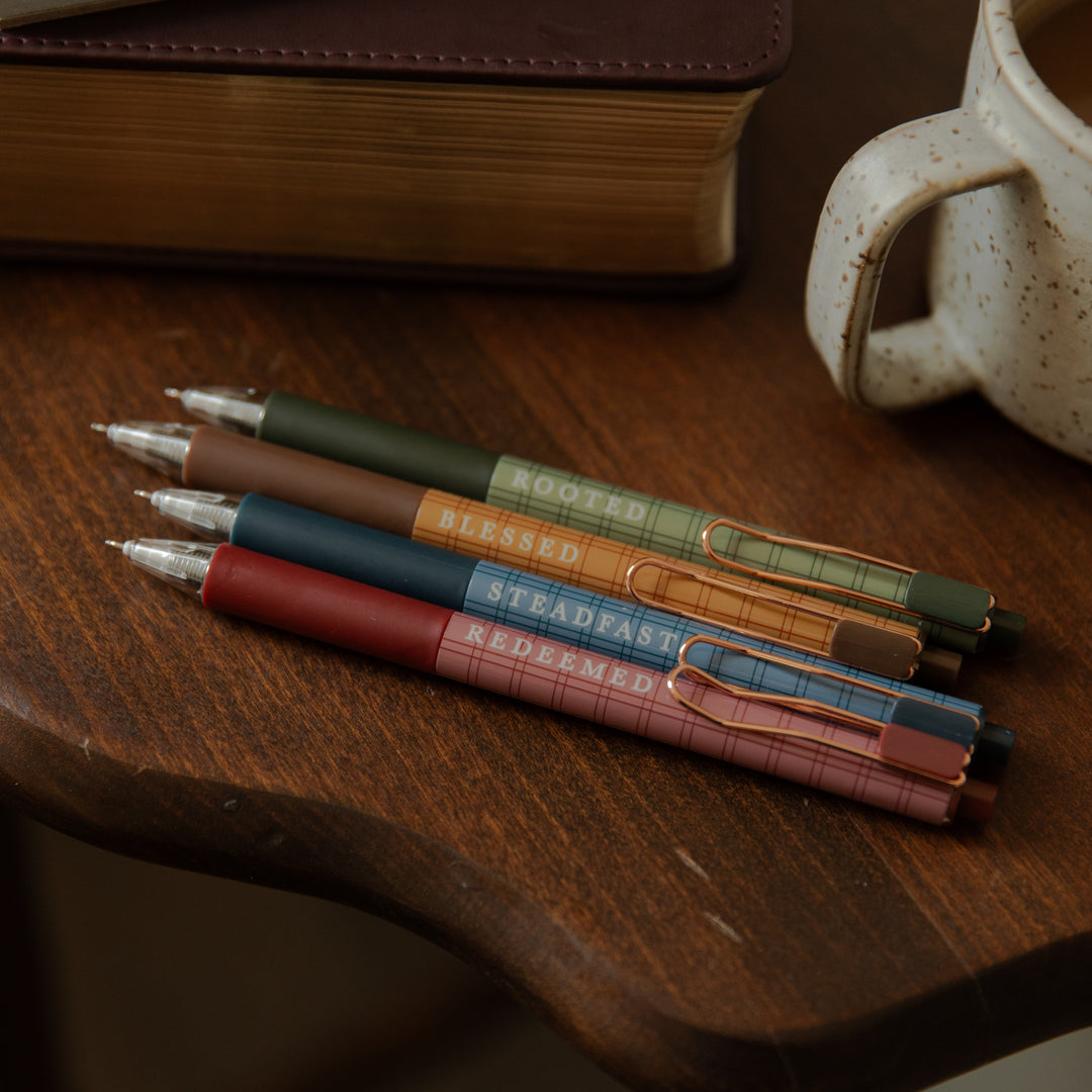 Four slim retractable pens in muted earthy colors resting on a wooden tabletop, with a book and a speckled mug partially visible in the background. The pens have plaid bands and printed words including 'ROOTED', 'BLESSED', 'STEADFAST', and 'REDEEMED'.