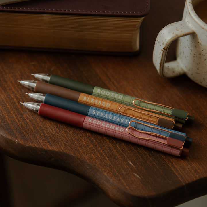 Four slim retractable pens in muted earthy colors resting on a wooden tabletop, with a book and a speckled mug partially visible in the background. The pens have plaid bands and printed words including 'ROOTED', 'BLESSED', 'STEADFAST', and 'REDEEMED'.