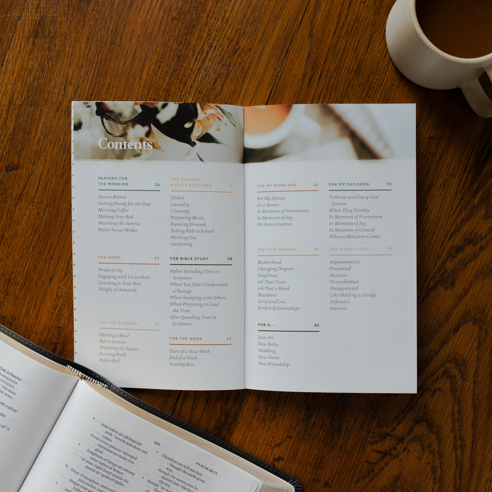 The Prayers for your Day opened to the table of contents page on a wooden table near a coffee cup