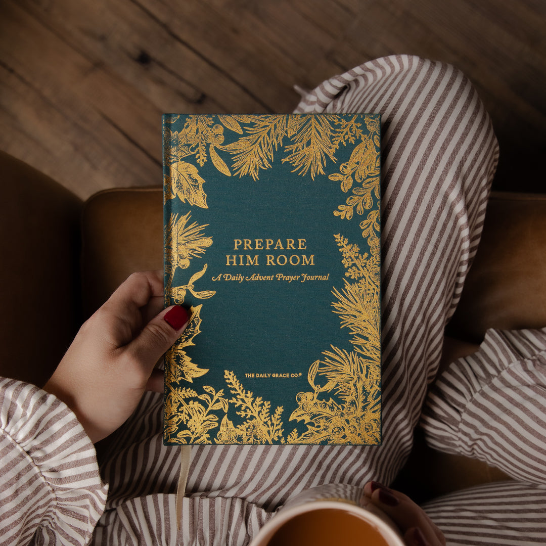A person in striped pajamas holds a teal journal with ornate gold botanical borders and the title 'PREPARE HIM ROOM' on the cover, photographed in a cozy indoor setting with a wooden floor and chair visible.