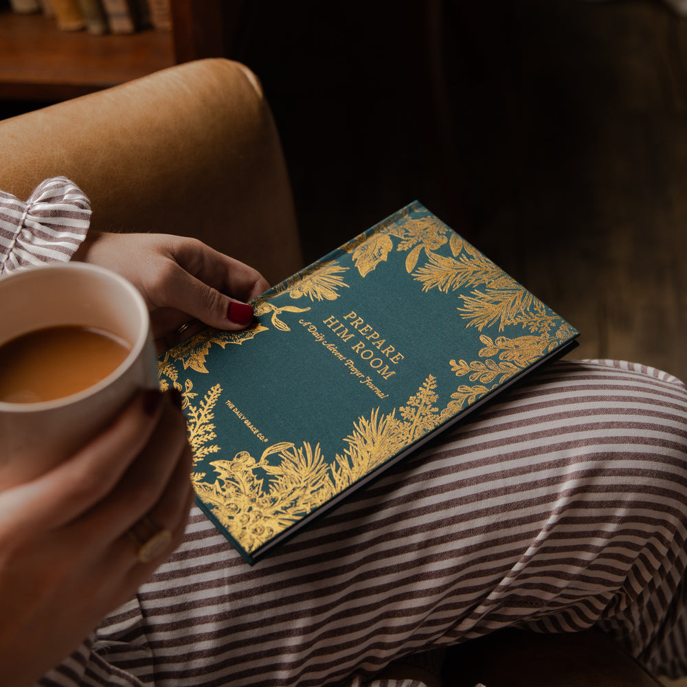 A person in striped pajamas holds a white mug while resting a teal and gold decorative book or journal on their lap. The cover reads “Prepare the Hill Room” and features ornate gold botanical illustrations.
