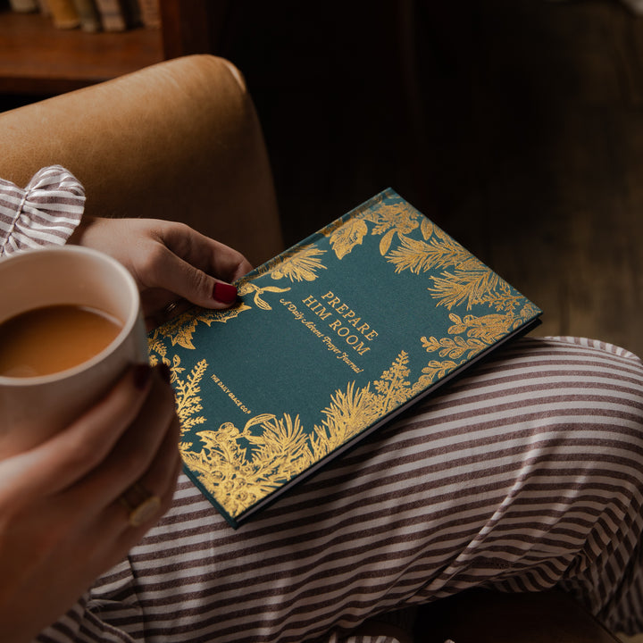 A person in striped pajamas holds a white mug while resting a teal and gold decorative book or journal on their lap. The cover reads “Prepare the Hill Room” and features ornate gold botanical illustrations.